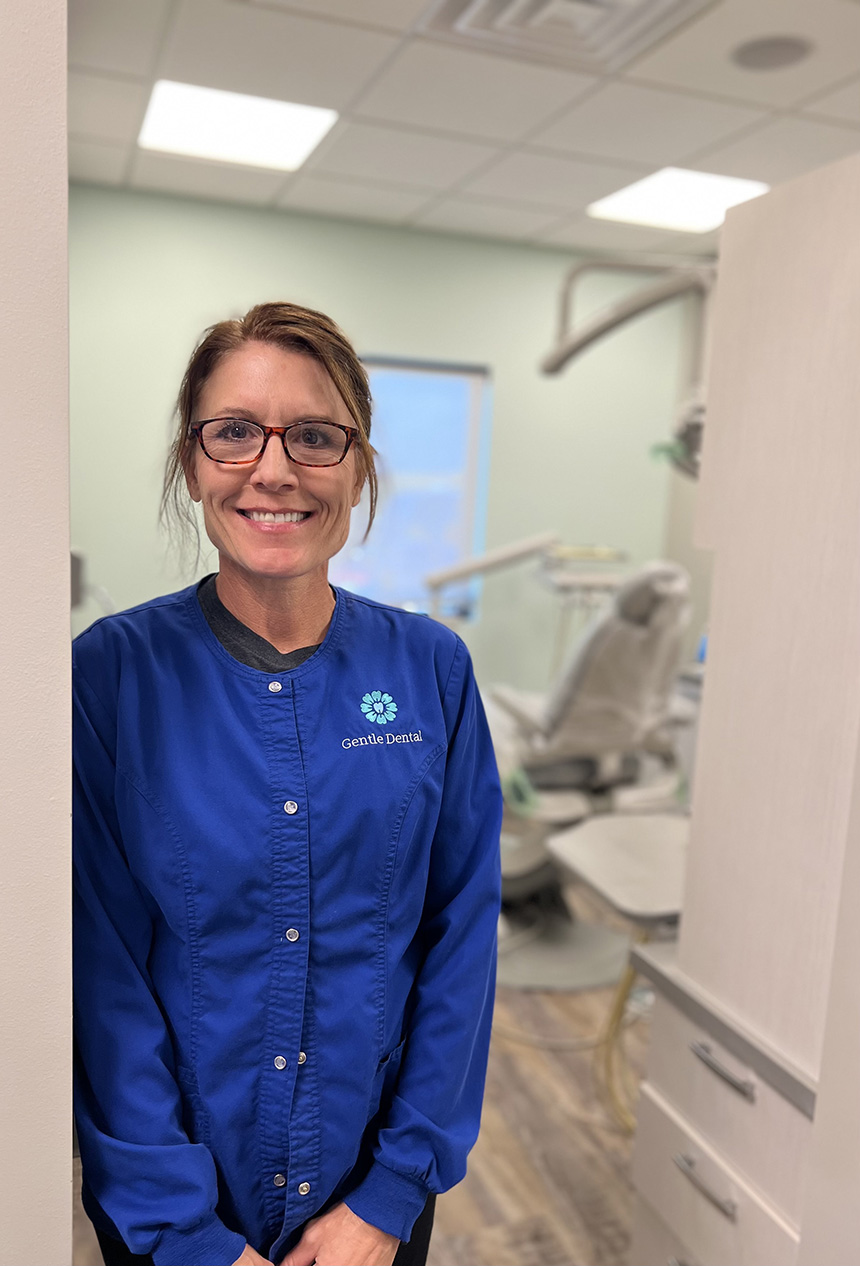 A woman wearing glasses stands in front of a dental office with a blue coat and a name tag, smiling at the camera.