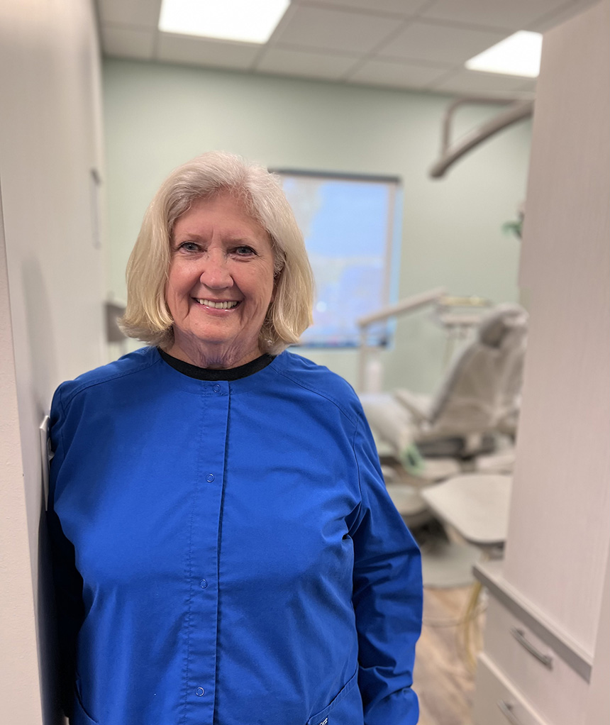 The image shows an older woman standing inside a dental office, wearing a blue shirt and white apron, with dental equipment visible in the background.